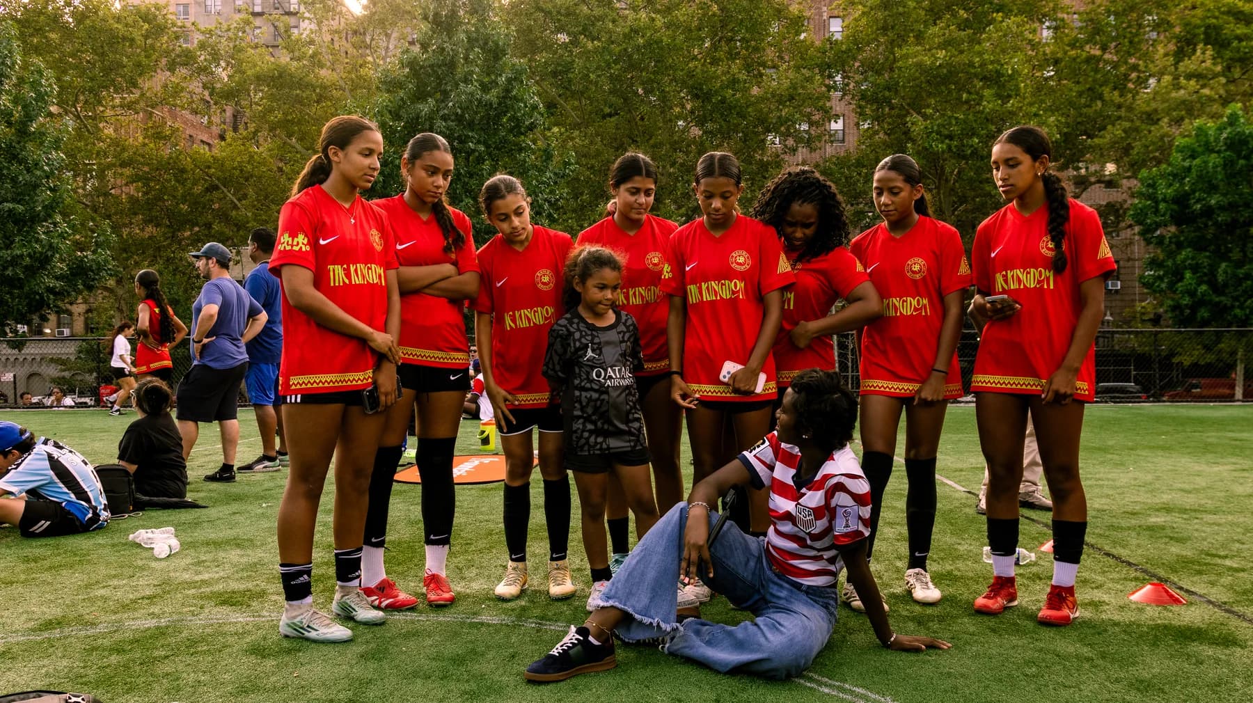 Girls soccer players in red Bamba Sports kits gathering on a New York field.