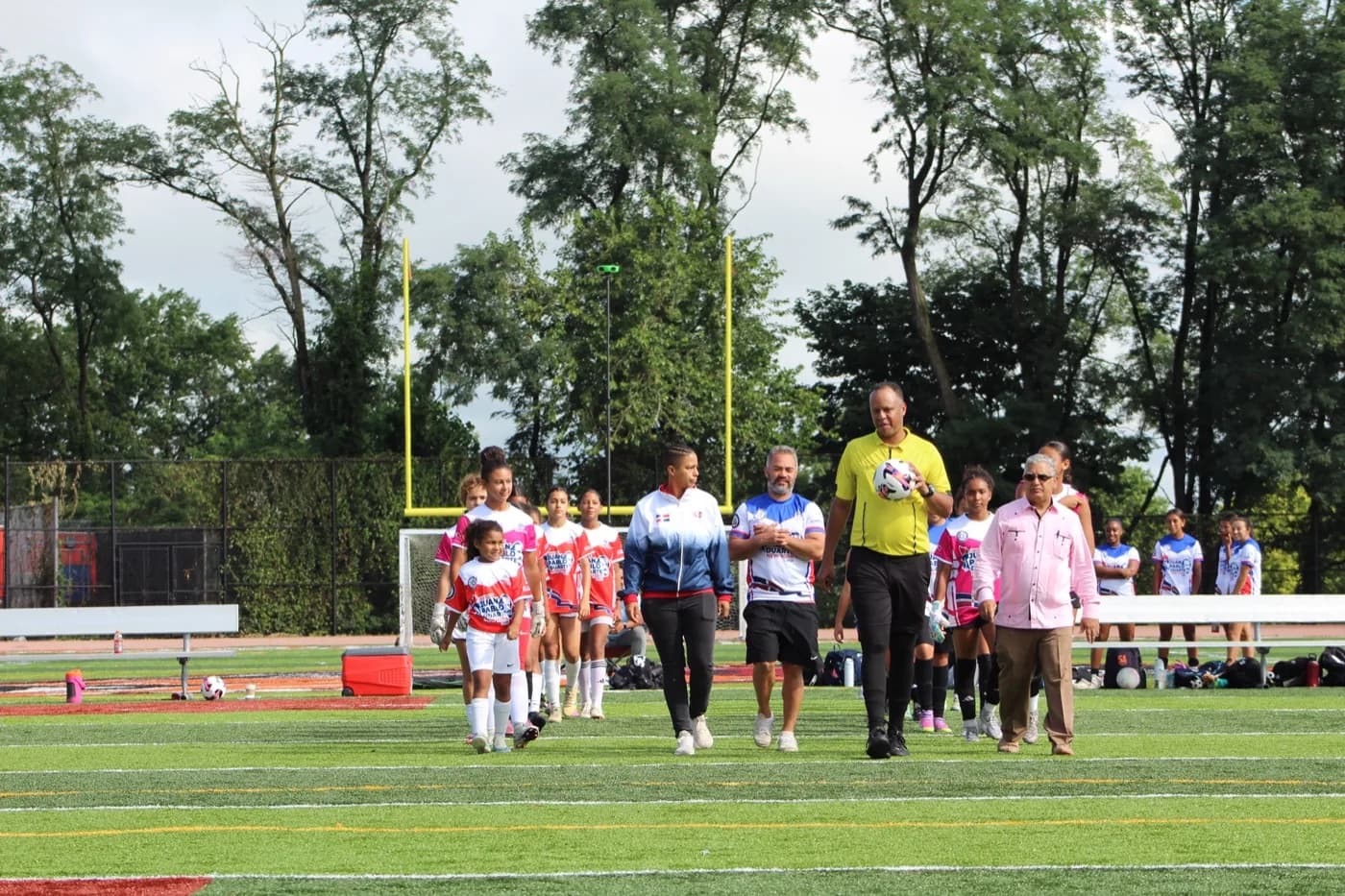 The referee carries the match ball as a guest coach, officials, and both squads walk out onto the field.